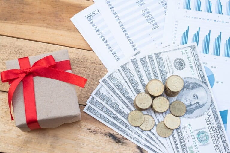 Coins, banknotes and spending statements are stacked next to a red bow paper gift box placed on a wooden table Coins, banknotes and spending statements are stacked next to a red bow paper gift box placed on a wooden table