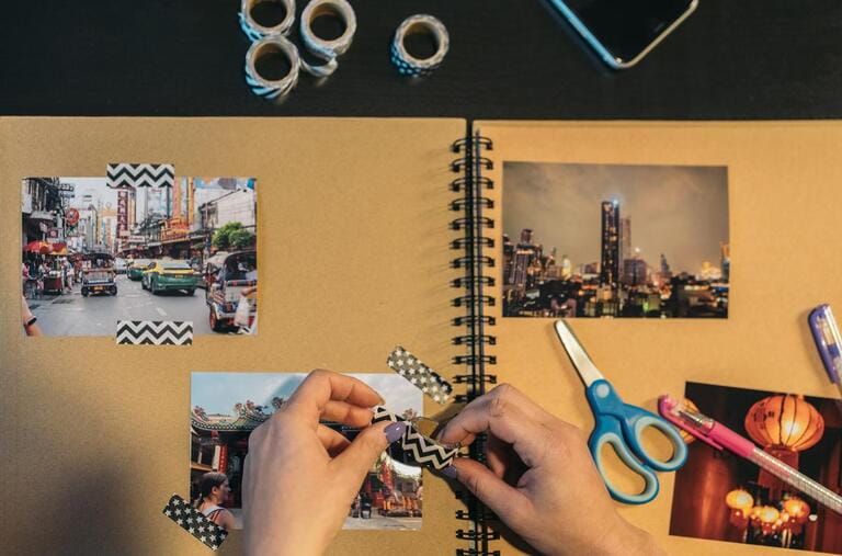 Image from above of the hands of an unrecognizable middle-aged woman picking up a piece of washi tape to make a handmade kraft travel album. Contains the word market in Thai.