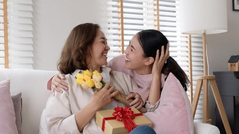 Attractive beautiful asian middle age mum sit with grown up daughter give gift box and flower in family moment celebrate mother day. Overjoy bonding cheerful kid embrace relationship with retired mom