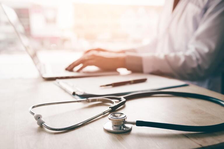 Stethoscope and background doctor using laptop at desk in clinic working on computer in office room.