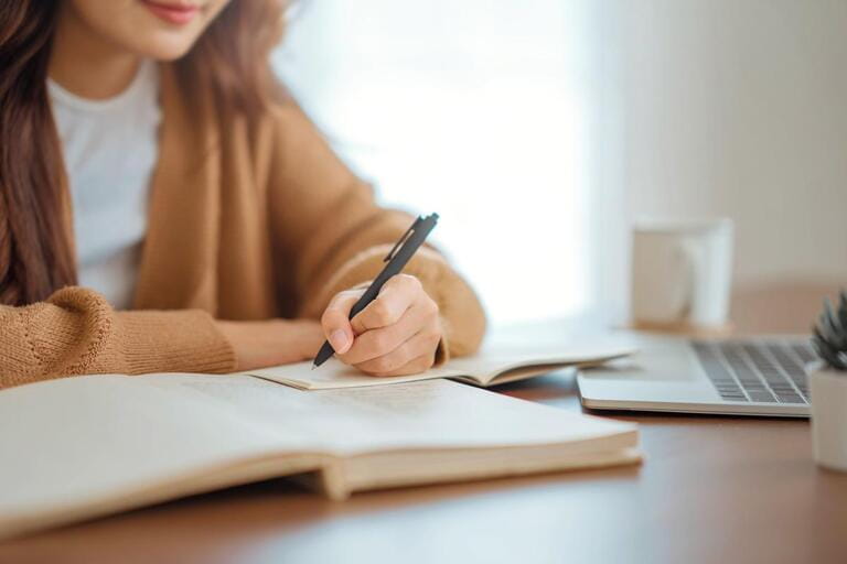 Closeup - Hand of woman writing in spiral notepad placed on wooden desktop with various items at home.