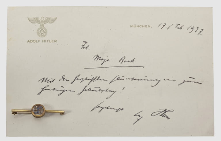 Maja Beck - a presentation brooch and birthday card from Adolf Hitler 1937,Gold brooch with pearl- and sapphire studded swastika in a circular,perforate filigreed bezel on a transverse bar with pin.Mark of fineness "585",length 48 mm,weight 2.6 g.The brooch pinned to a card with a raised national eagle in gold(darkened)above "Adolf Hitler" and the hand-written best wishes text "Frl.Maja Beck - Mit den herzlichsten Glückwünschen zum heutigen Geburtstag! Ergebenst Adolf Hitler"(Miss Maja Beck - With the most heartfelt best wishes on your birthday today!,Additional-Rights-Clearences-Not Available